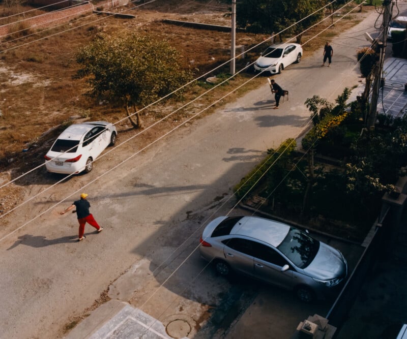 Aerial view of a quiet street with three parked cars, a man in red pants walking, and two people walking a dog. Trees and greenery are visible alongside the road.