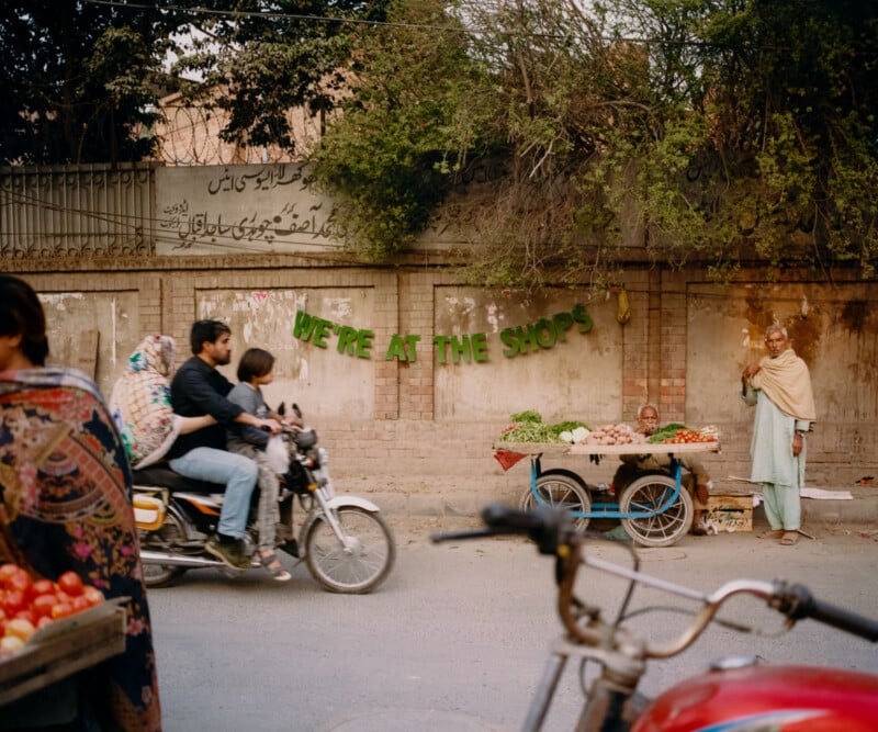 A busy street scene shows a family riding a motorcycle past a fruit and vegetable vendor with a cart. Green letters on the wall behind the cart spell “WE’RE AT THE SHOPS.” A woman stands at the vendor’s cart.