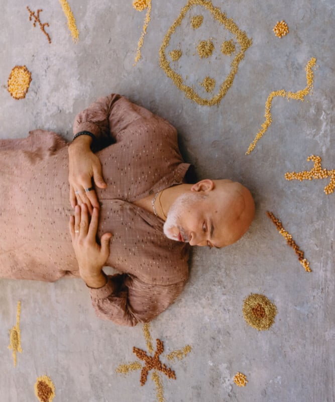 A bald man with a white beard lies on his back on a concrete floor, wearing a brown patterned shirt. Around him, various grains or seeds are arranged in abstract shapes and patterns on the ground.