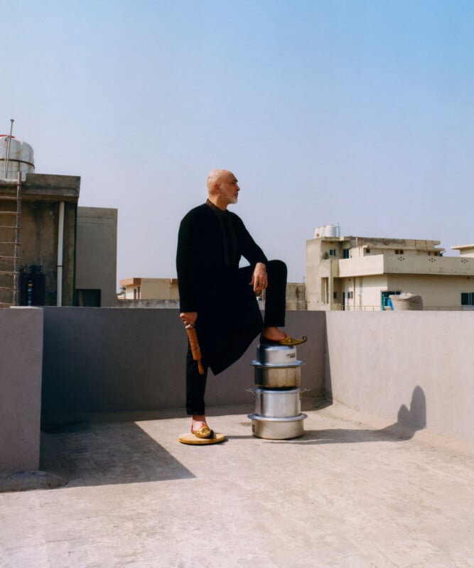A man in traditional black attire stands confidently on a rooftop with one foot on a stack of metal pots, holding a wooden rolling pin and wearing yellow slippers, under a clear blue sky.