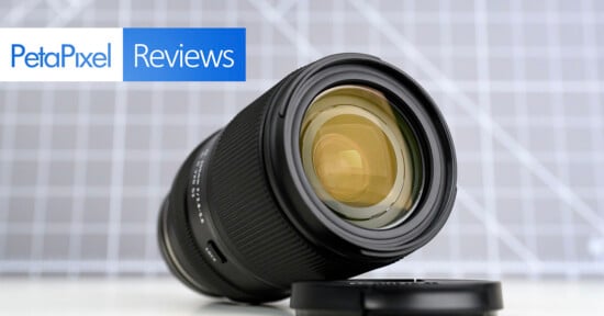 A camera lens rests on its side next to a lens cap, with a Petapixel Reviews logo in the corner and a grid-patterned background.