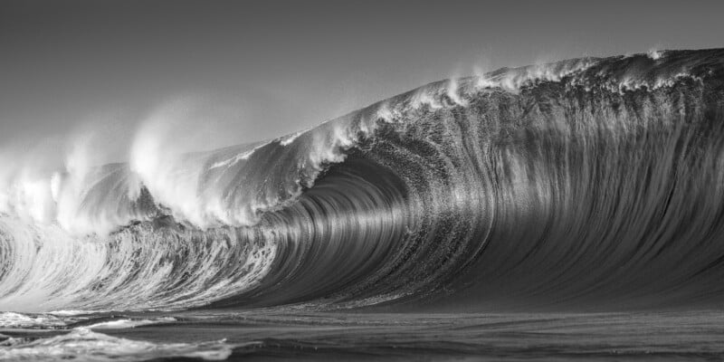 A dramatic black-and-white photo of a large ocean wave curling and crashing, with mist spraying off the crest and textured water patterns visible throughout the wave.