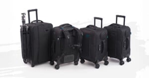 Four black rolling suitcases stand side by side on a white background; one has a tripod attached, and another is shown with backpack straps visible.
