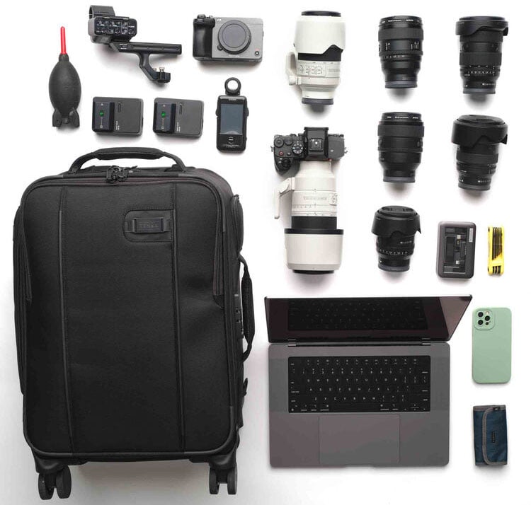 Una maleta, una cámara, varias lentes de cámara, una computadora portátil, un teléfono inteligente, una billetera, un cargador de batería, baterías, un soplador y accesorios de cámara cuidadosamente dispuestos sobre una superficie blanca.