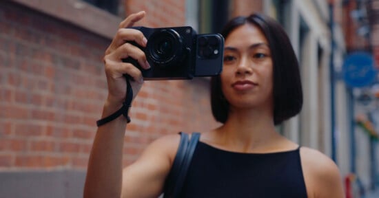 A person with short dark hair holds a camera with a smartphone attached, standing outdoors on a city street with brick buildings in the background.