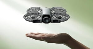 A small drone with protective guards hovers above an outstretched hand against a light green background.
