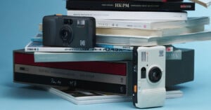 Two vintage-style Kodak cameras, one black and one white, are displayed in front of a stack of books and magazines against a light blue background.