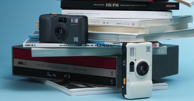 Two vintage-style Kodak cameras, one black and one white, are displayed in front of a stack of books and magazines against a light blue background.