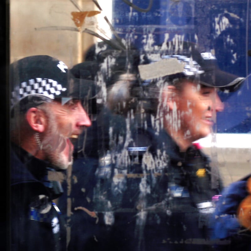 Two police officers in uniform, partially obscured by a dirty, scratched glass surface, stand side by side. Their faces are blurred and distorted by the marks on the glass.