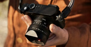 A person in a brown shirt holds a black digital camera with a wide lens and a braided strap, adjusting the settings with their hand.