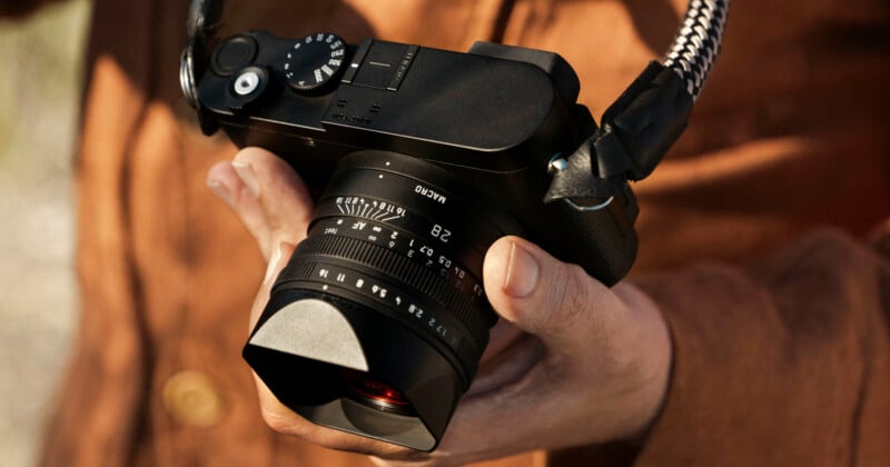 A person in a brown shirt holds a black digital camera with a wide lens and a braided strap, adjusting the settings with their hand.