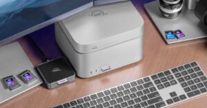 A desktop setup with a wireless keyboard, external hard drives, a camera lens, several SD cards, and a computer monitor on a wooden desk. The devices are neatly arranged beside each other.