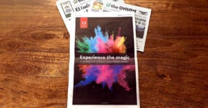 A newspaper on a wooden table features a colorful explosion of powder and the text, “Experience the magic of buying new software, every month, forever.” Other newspapers are partially visible underneath.