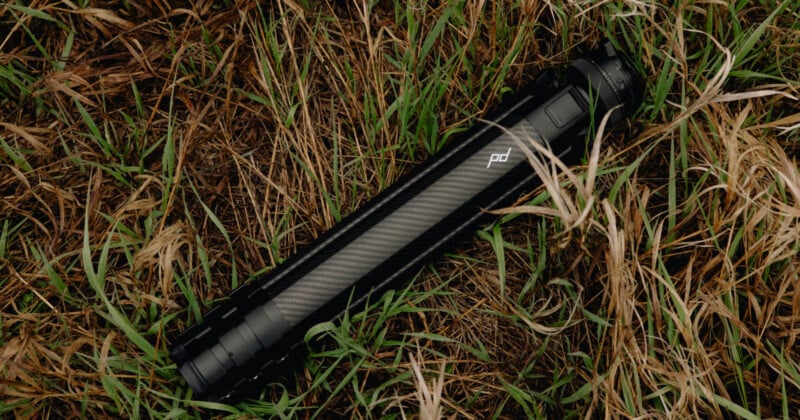A black, collapsible camera tripod with carbon fiber legs lies folded on dry, patchy grass.