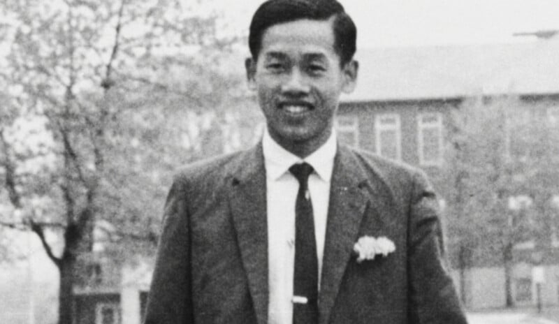 A black-and-white photo of a man in a suit and tie, smiling outdoors with trees and a building in the blurry background.