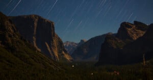 A nighttime landscape of a valley surrounded by rugged mountains, featuring long, bright star trails streaking across the sky above dark forests and dramatic rock formations.