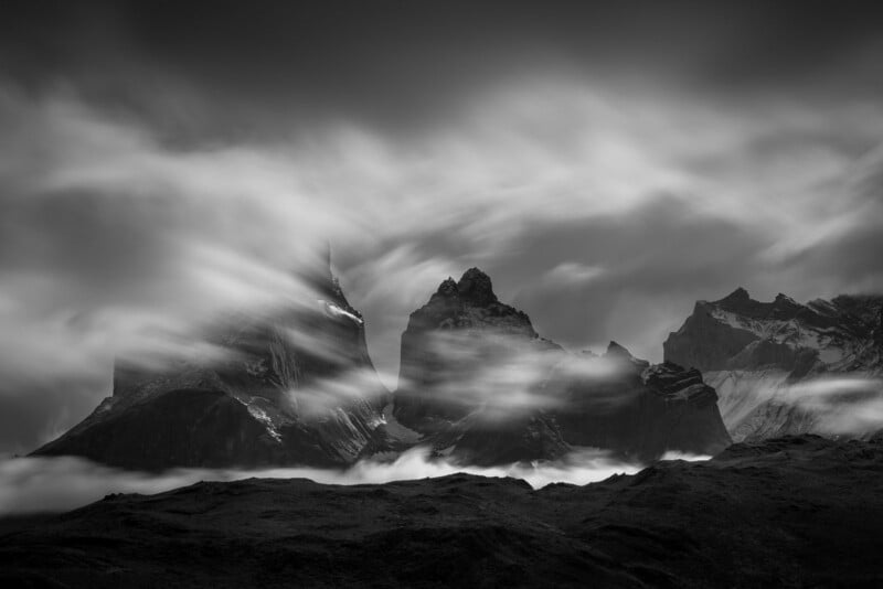Black and white photo of jagged mountain peaks shrouded in flowing clouds, creating a dramatic and mysterious atmosphere with strong contrasts and soft, misty streaks through the scene.