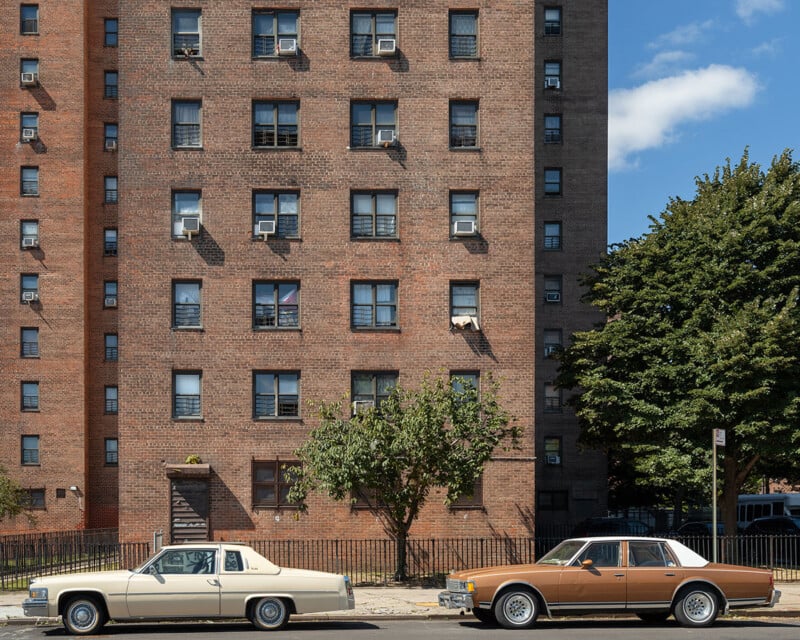 Dos coches antiguos, uno color crema y otro marrón, estaban aparcados en la calle, frente a un alto edificio de apartamentos de ladrillo con muchas ventanas. También puedes ver árboles y cielo azul y nubes blancas.