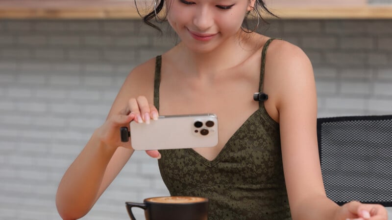 Una mujer con un vestido verde sostiene un teléfono inteligente y toma una foto de un café con leche en una taza negra con una sonrisa amable. Está sentada en una mesa con una pared de ladrillos claros como telón de fondo.