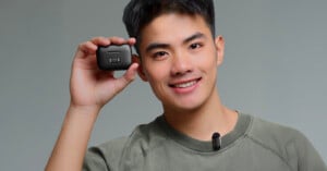 A smiling person wearing a green shirt holds a small black wireless device next to their face. A clip-on microphone is attached to their shirt collar. The background is plain gray.