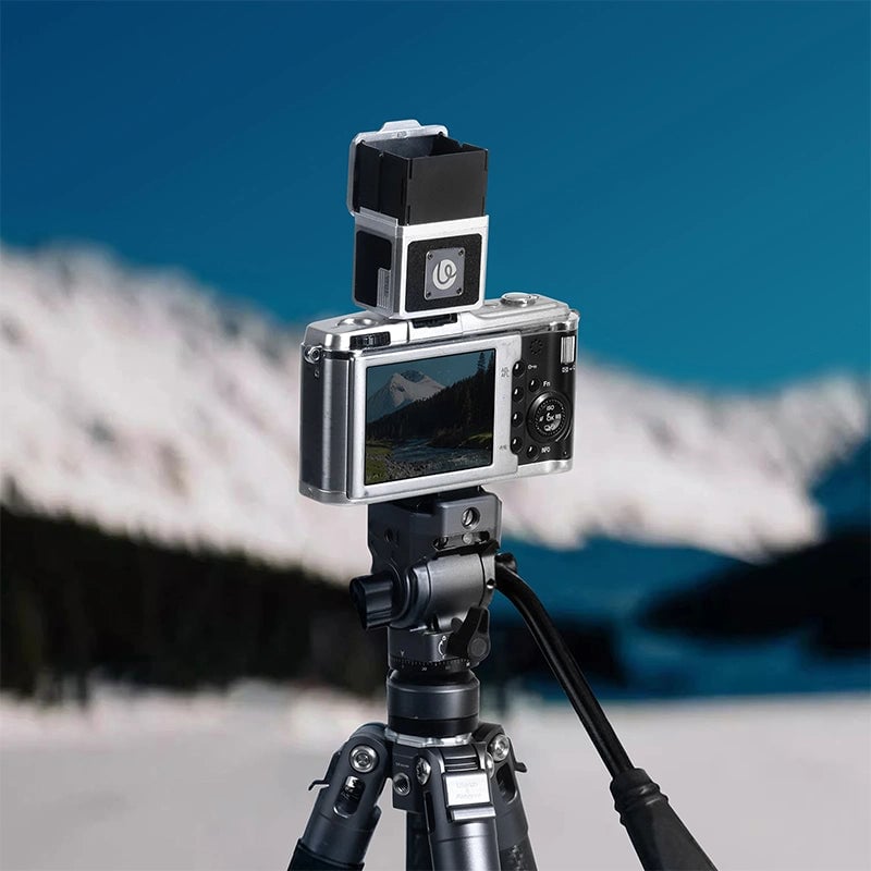 A digital camera mounted on a tripod is positioned outdoors, with a small external device attached on top. Snow-covered mountains and a blue sky are visible in the blurred background.