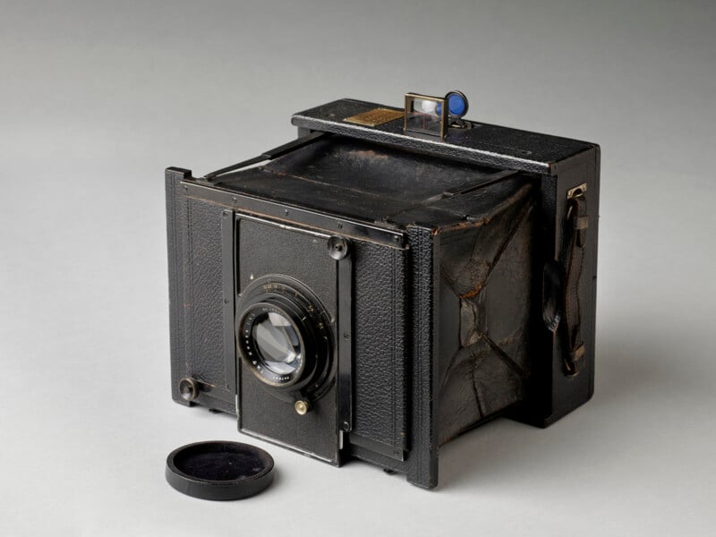 A vintage black box camera with a bellows body, lens in front, and a lens cap lying nearby, displayed on a plain light surface. The camera shows signs of wear and includes a side leather strap.
