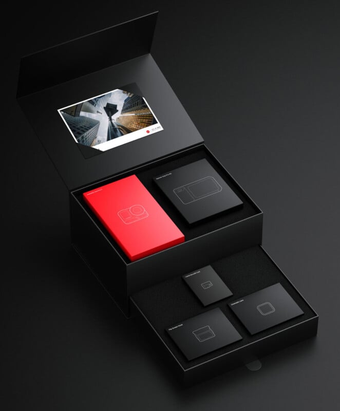 Una elegante caja negra, con la tapa abierta, revela varios elementos cuidadosamente ordenados: un folleto rojo, un folleto negro con los contornos gráficos de una cámara y un teléfono móvil, y una impresión fotográfica, todo en un estilo moderno y minimalista sobre una superficie oscura.