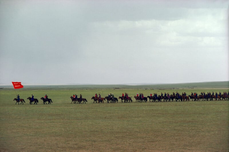 A group of people on horseback ride across a vast, open grassland. One rider at the front carries a large red flag. The sky is overcast and the landscape is flat and expansive.