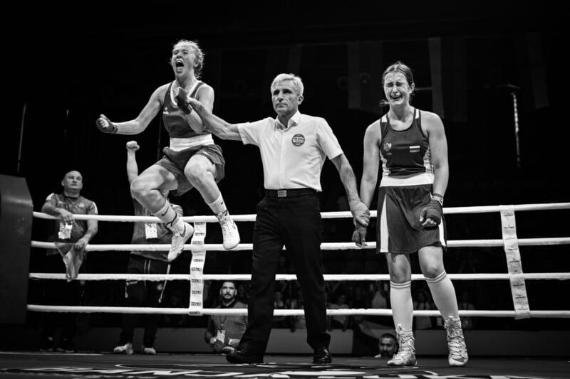 A referee stands in a boxing ring holding the arms of two female boxers. One boxer jumps joyfully, celebrating victory, while the other stands with tears and disappointment. The crowd watches in the background.
