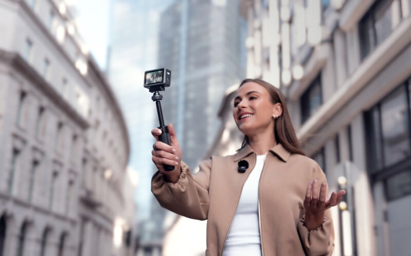 A woman smiles and talks while holding a camera on a selfie stick, recording herself on a city street with tall modern and classical buildings in the background.