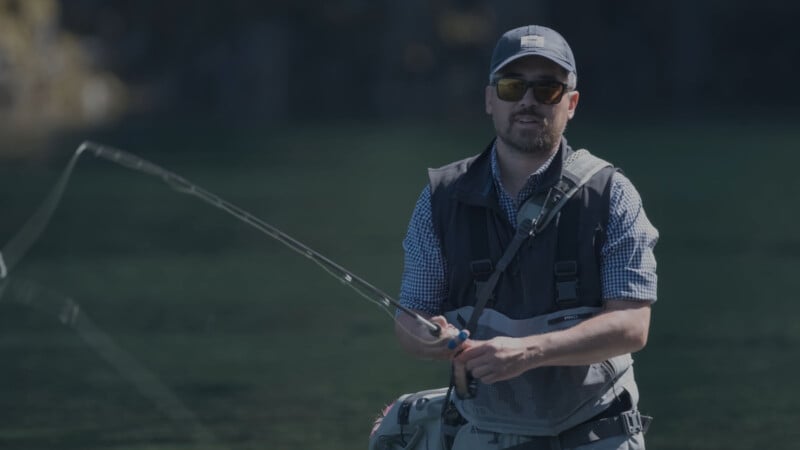 A man wearing sunglasses, a cap, and a fishing vest stands outdoors by the water, holding a fishing rod and casting a line.