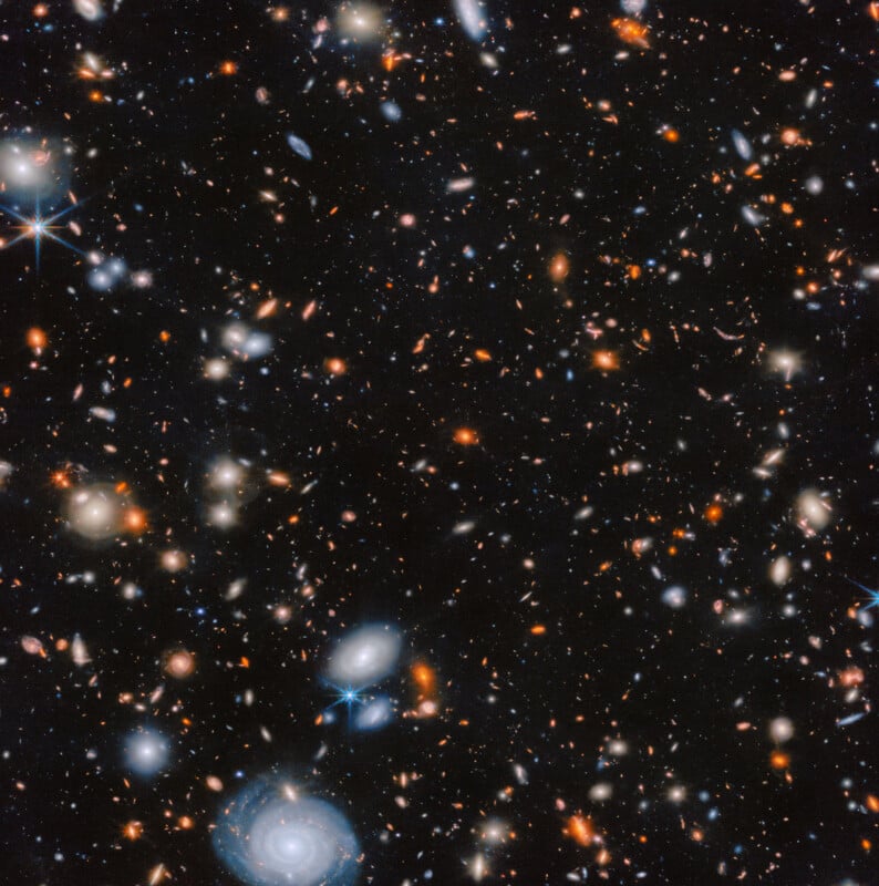 A deep-space image showing countless distant galaxies in various shapes and colors, with bright spirals, elliptical forms, and glowing stars scattered across a dark background.