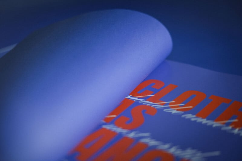 A close-up of a blue page being turned in a book, revealing bold red and white text underneath, partially reading "CLOTH IS" against the blue background. The image is softly focused and brightly lit.