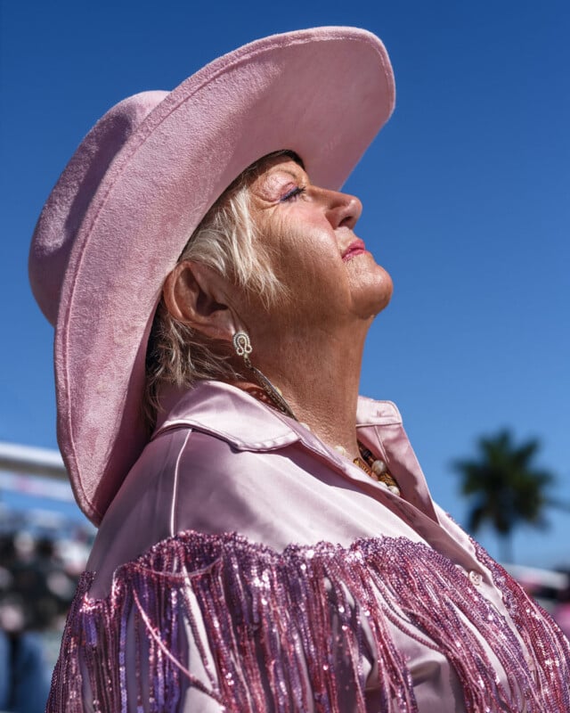 Una anciana que lleva una chaqueta rosa con flecos y un sombrero de ala ancha se encuentra bajo un cielo azul claro con los ojos cerrados y el rostro mirando al sol; al fondo hay una palmera borrosa.