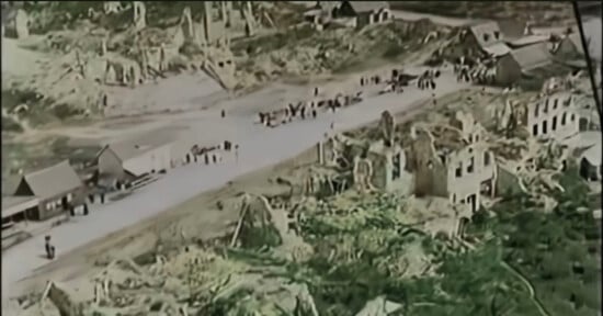 Aerial view of a street lined with people amid the ruins of destroyed buildings and debris, suggesting devastation in a war-torn area.
