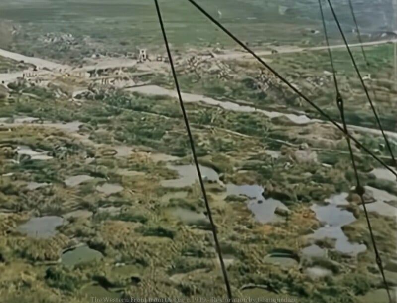 Una vista aérea de un campo de batalla de la Primera Guerra Mundial, que muestra un paisaje plagado de cráteres, trincheras y escasa vegetación. Los cables eléctricos cruzan el primer plano y los edificios distantes son visibles cerca del horizonte.