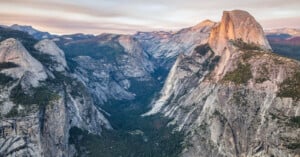A dramatic view of Yosemite Valley with steep granite cliffs, dense forest below, and the iconic Half Dome rising prominently under a soft, pastel sky at sunset.