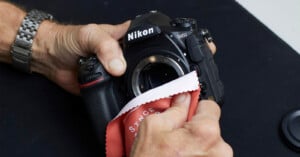 A person uses a red microfiber cloth to clean the sensor area of a Nikon DSLR camera, holding the camera in one hand and wiping with the other. The camera lens has been removed.