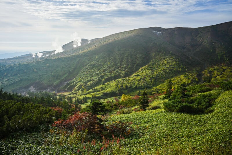 Lush green hills with scattered trees and patches of red foliage, emitting plumes of steam, under a partly cloudy sky.