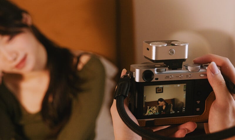 A close-up of hands holding a camera, focusing on a woman with long dark hair and a green top, who is sitting and posing in a softly lit indoor setting; her image is visible on the camera screen.