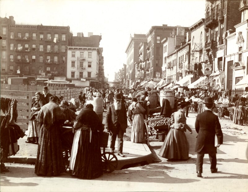 Una fotografía en tonos sepia muestra una concurrida calle de la ciudad a principios del siglo XX, con gente caminando con trajes de época, comprando en carritos de vendedores y socializando entre edificios altos y puestos de mercado.
