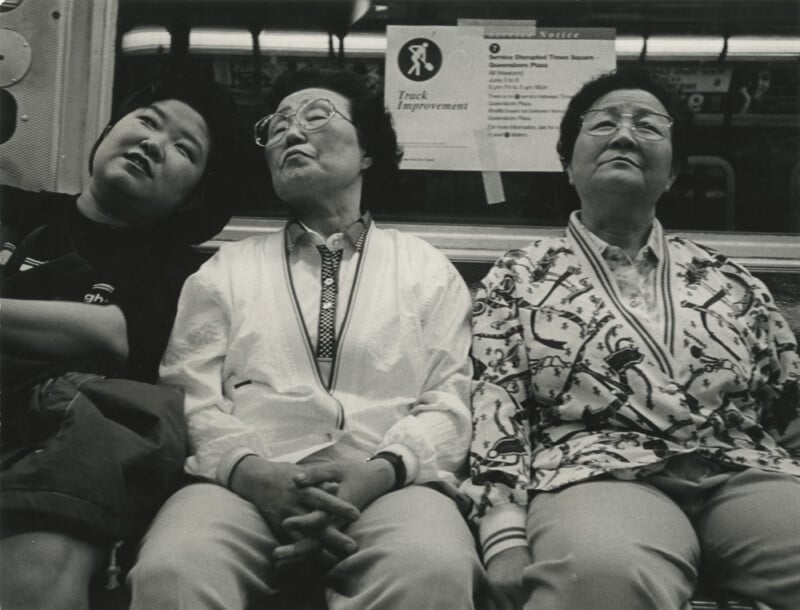 Tres ancianas con gafas sentadas muy juntas en un banco del metro con expresiones neutras o cansadas. uno "seguimiento de mejoras" Se colocaron avisos en la pared detrás de ellos. Las fotos están en blanco y negro.