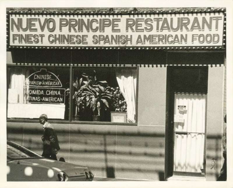 Fotografía en blanco y negro de un restaurante con eslogan. "NUEVO Restaurante PrÍncipe: La más deliciosa cocina china-española-americana." Un hombre pasó junto a la ventana y un segundo cartel indicaba la comida en chino, español e inglés.