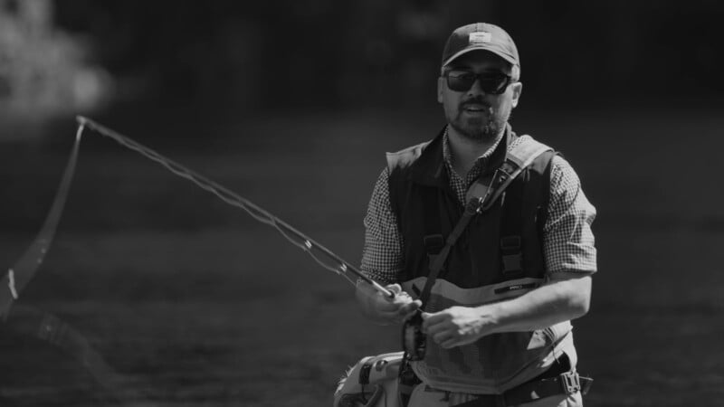 A man wearing sunglasses, a cap, and a fishing vest stands in shallow water while fly fishing, holding a fishing rod mid-cast. The background is out of focus, with natural light highlighting the scene.