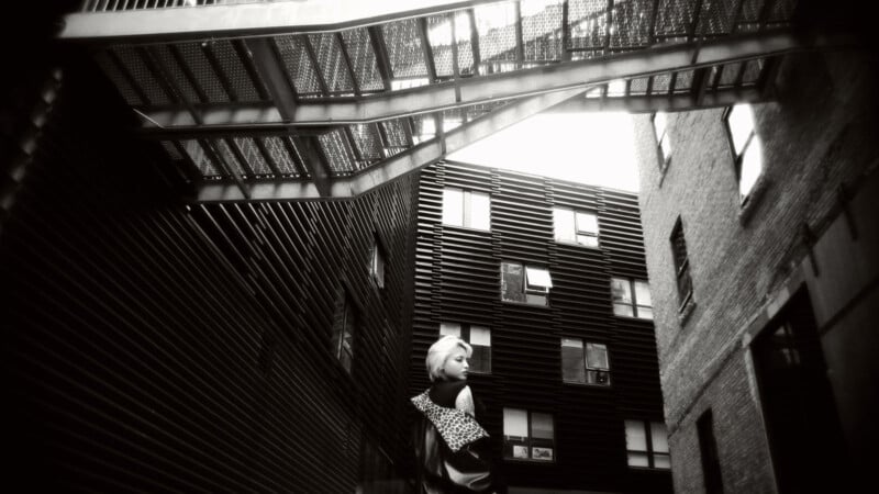 Una fotografía en blanco y negro de un hombre parado en un callejón estrecho entre edificios modernos, mirando hacia atrás. Una escalera de metal cuelga encima y las ventanas rectangulares reflejan la luz desde arriba.