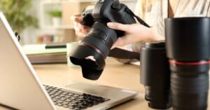 A person holds a DSLR camera in front of an open laptop on a desk, with additional camera lenses nearby. The scene suggests photography work or photo editing.