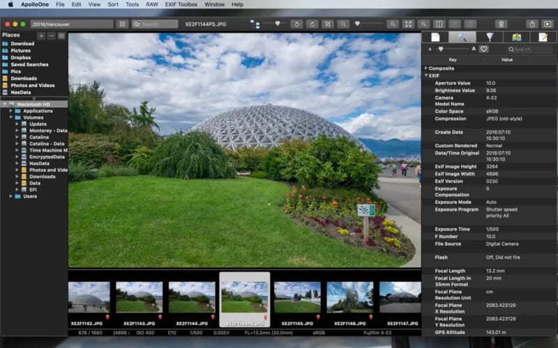 A large glass-domed conservatory stands behind a green lawn and colorful flower beds under a partly cloudy sky, with people walking on a nearby path.