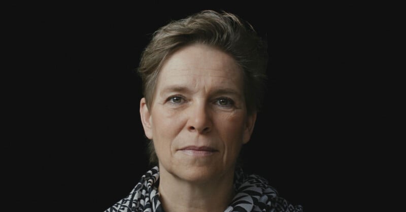 A middle-aged woman with short hair and a calm expression looks directly at the camera. She wears a black and white patterned scarf and is set against a plain black background.