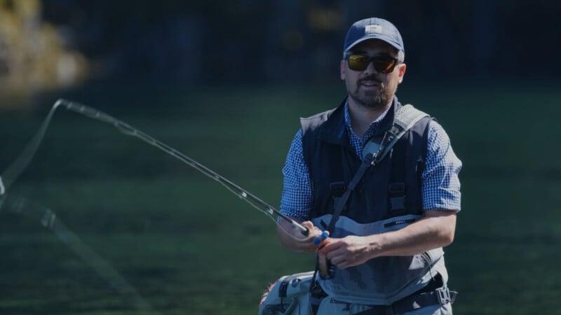 A man wearing sunglasses, a cap, and a fishing vest is standing outdoors, holding a fishing rod over clear green water, preparing to cast his line.