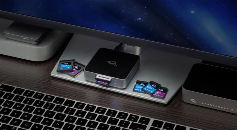 A computer desk setup with a keyboard, monitor, and several Thunderbolt 3 devices, including a memory card reader dock holding SD cards, all arranged neatly on a dark wood surface.